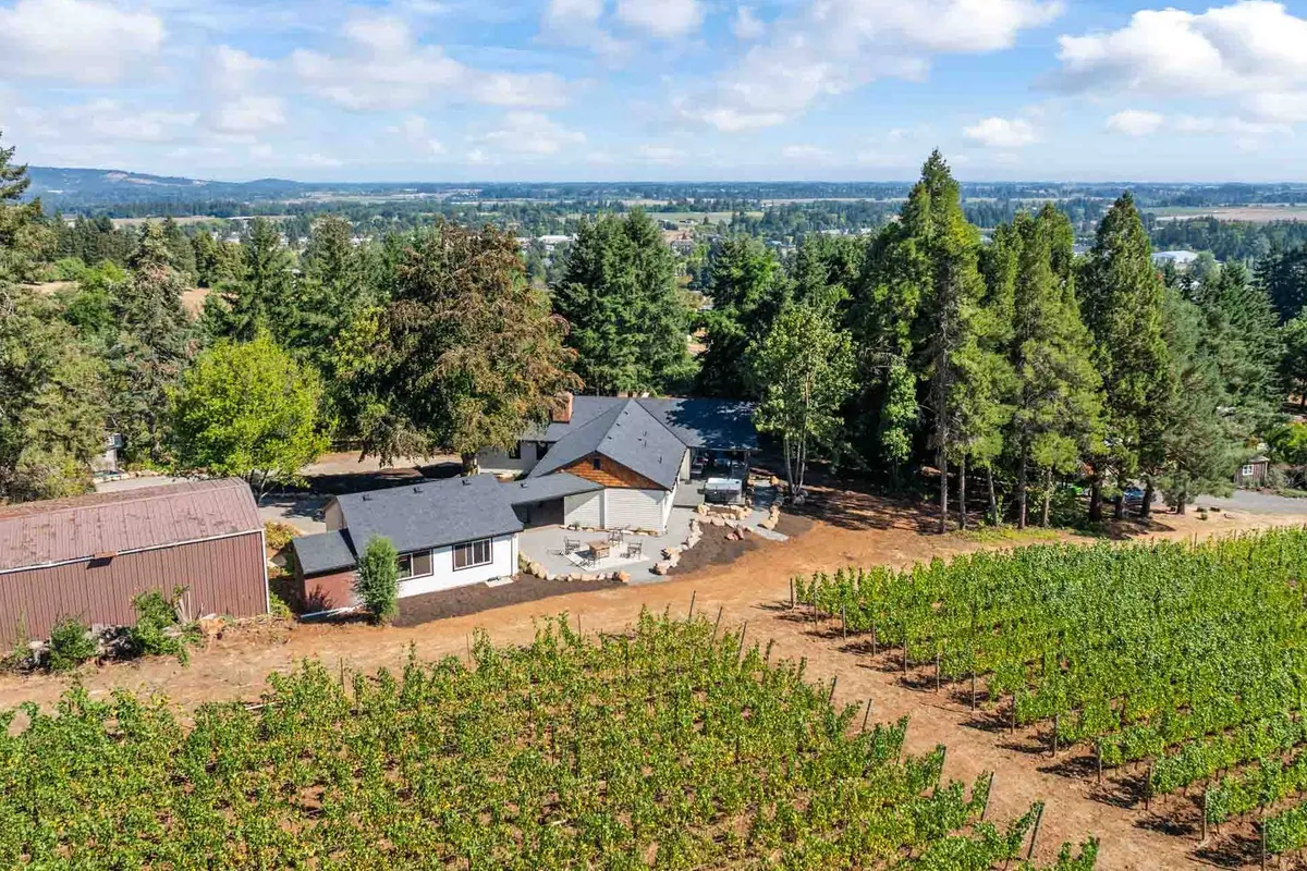 A sweeping exterior view of the house set against rows of lush vineyards, with cozy outdoor seating under the shade of mature trees. The scene captures the essence of the Dundee Hills wine country, with visible vineyard rows and hints of the home’s inviting patio area—ideal for wine tasting and gatherings.