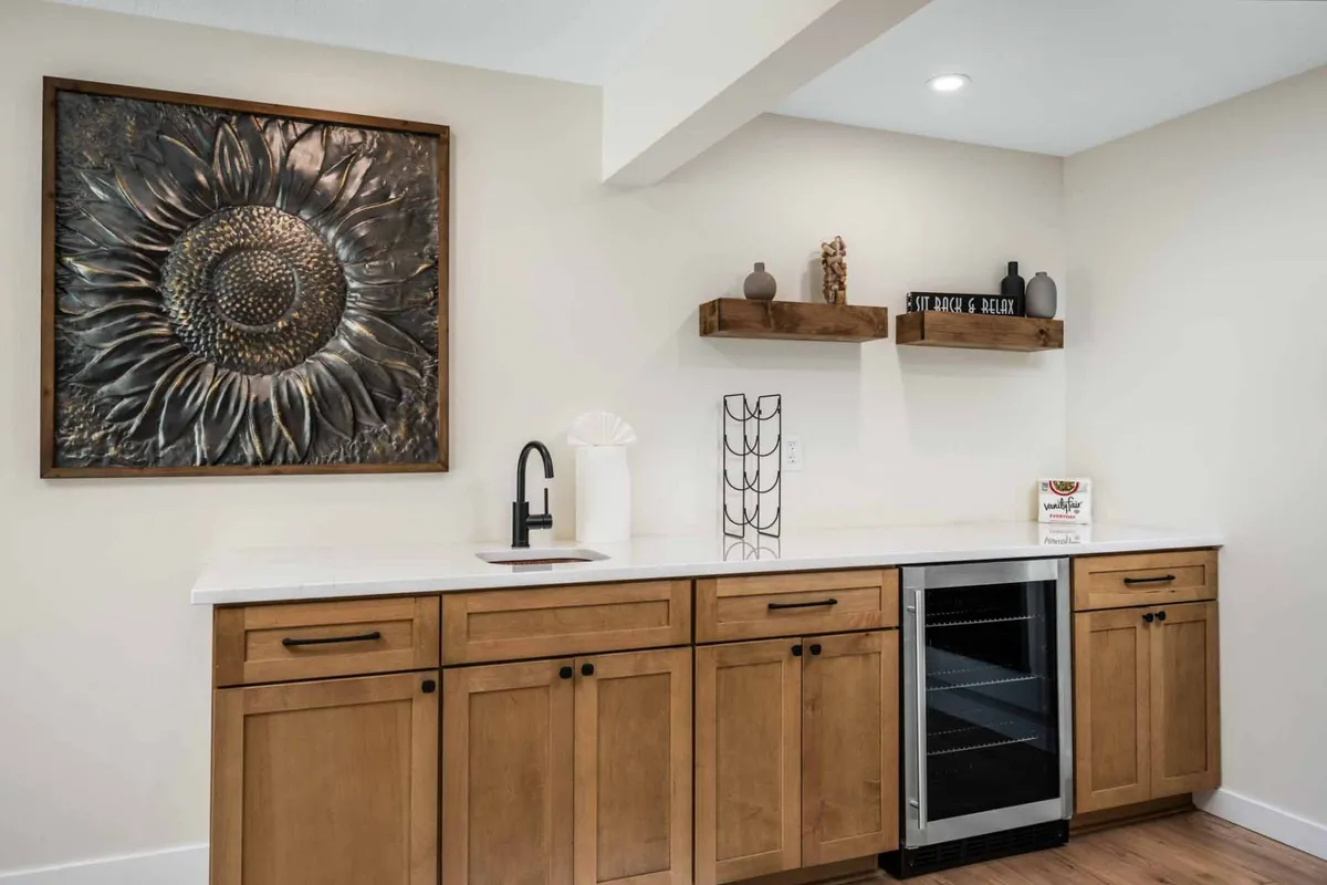 An inviting interior scene featuring a wine cooler, wooden shelves, and metal sunflower artwork, creating a warm and welcoming ambiance perfect for socializing. The cabinetry and wine accessories hint at a well-equipped wet bar area ideal for sharing toasts and celebrating together.