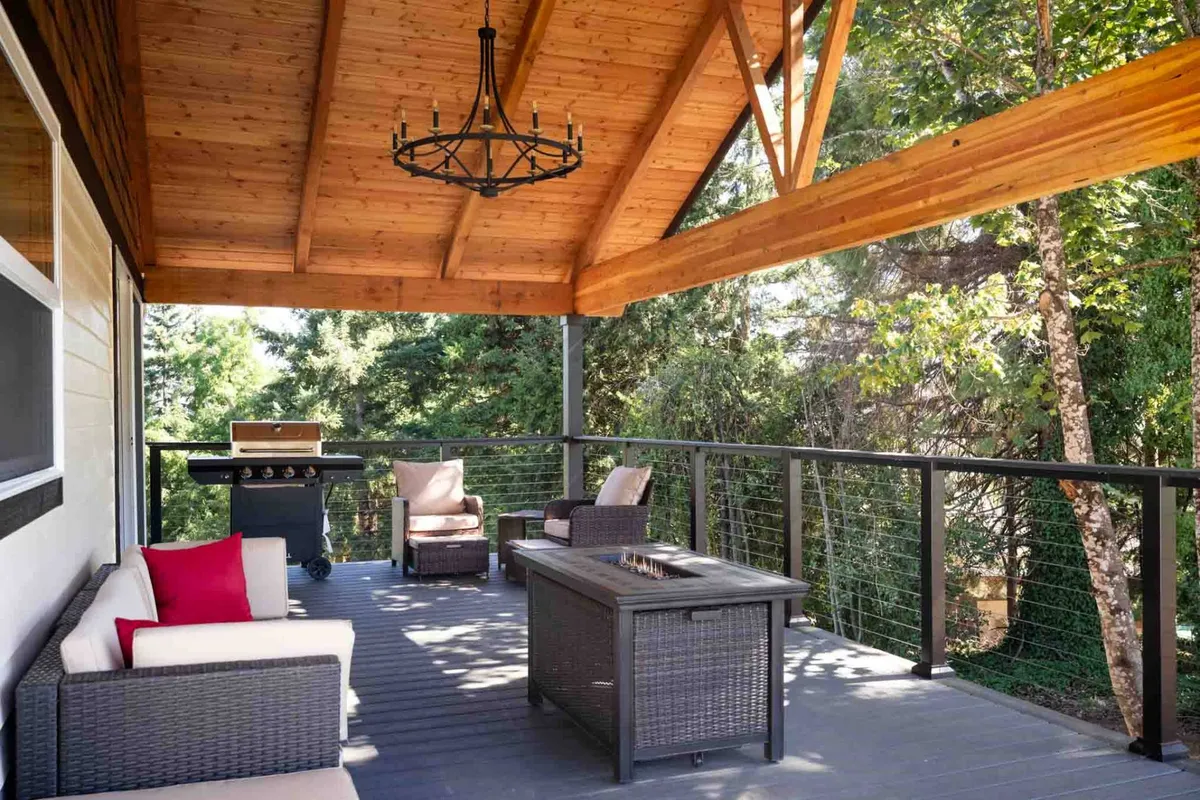 A cozy covered area of the deck is shown, featuring plush outdoor seating, a fire pit, a grill, and surrounding greenery. The wooden ceiling and intimate arrangement invite guests to relax, dine, and socialize in comfort while enjoying the outdoors.