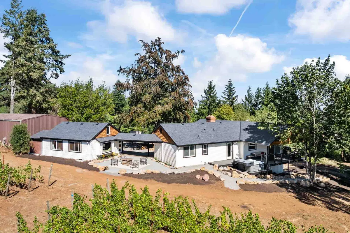 An inviting exterior view of Courtyard Cottage featuring a welcoming outdoor seating area surrounded by mature trees and a rolling vineyard just beyond. Comfortable chairs gather around a fire pit, with the home’s facade and greenery creating a secluded, peaceful setting. The lighting hints at golden hour, capturing the serenity of the rural landscape.