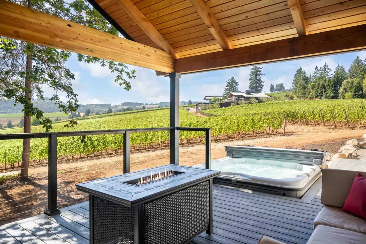 This image captures a sprawling deck area with a recessed hot tub, a propane fire pit, luxury lounge seating, and sweeping views of the surrounding vineyard. The wooden beams and ambient lighting create an inviting outdoor living space ideal for relaxation and gatherings.