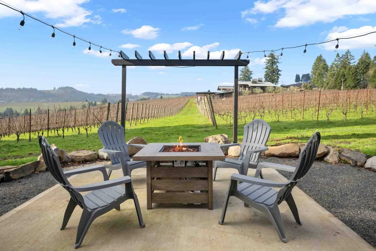 An intimate outdoor area featuring a fire pit surrounded by Adirondack chairs, all overlooking the picturesque vineyard. String lights add a festive touch, hinting at cozy evenings spent sharing stories and enjoying local wines by the fire as the sun sets over the vines.