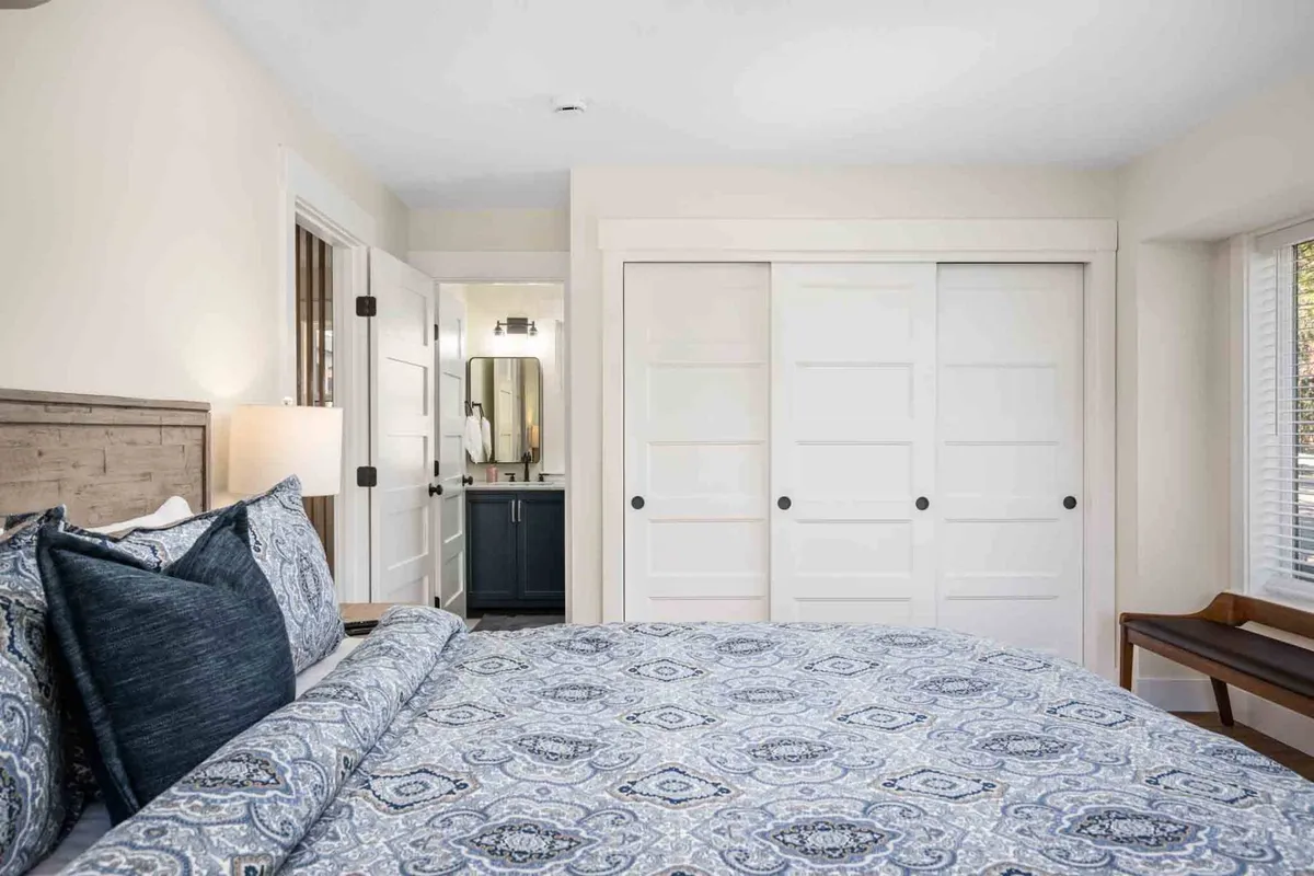 A cozy bedroom with patterned bedding, a nightstand and lamp, closet and bathroom doors, and a bench by the window. The design is modern and inviting, with plenty of space for guests and hints of convenient amenities.