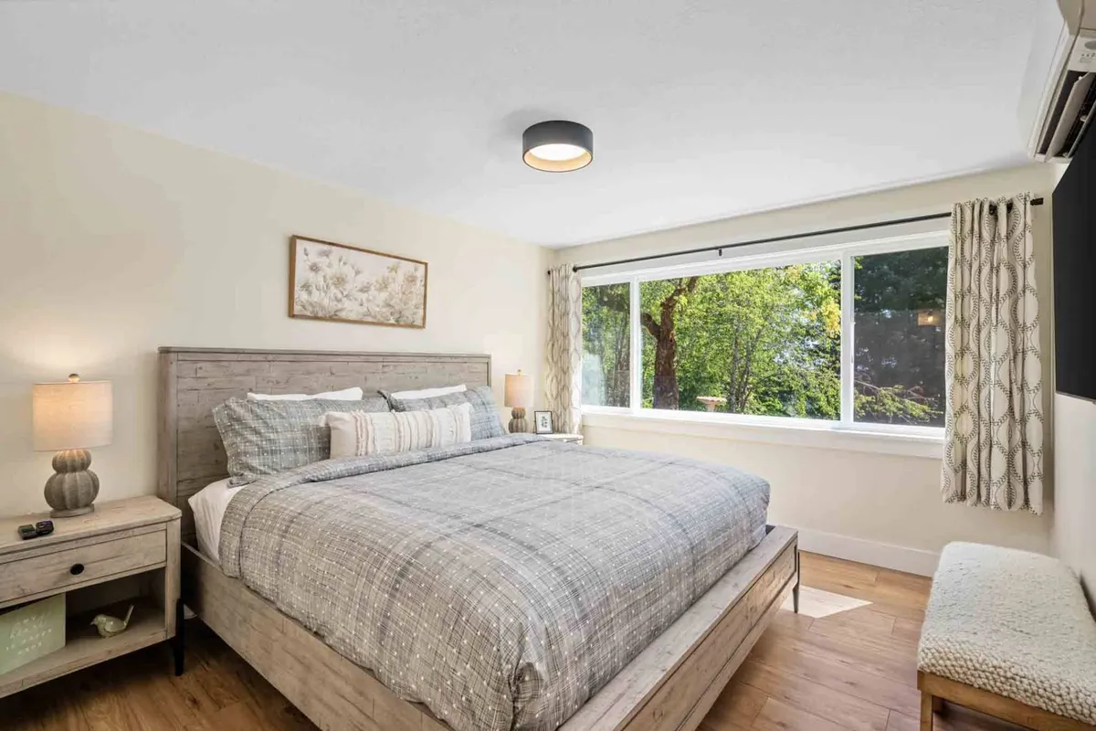 A tranquil bedroom featuring a comfortable bed with crisp white linens, a large window letting in natural light, a nightstand with a lamp, and minimalist decor. The room exudes a sense of calm and relaxation, ideal for unwinding after a day exploring the vineyards.