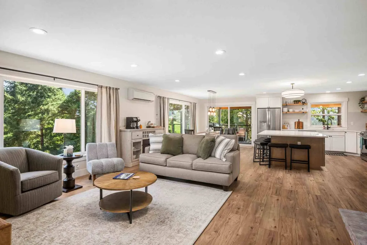 An open-concept great room featuring a comfortable sofa, modern coffee table, and a kitchen island visible in the background. The space is styled for both relaxation and socializing, with ample seating and natural light streaming in from large windows.