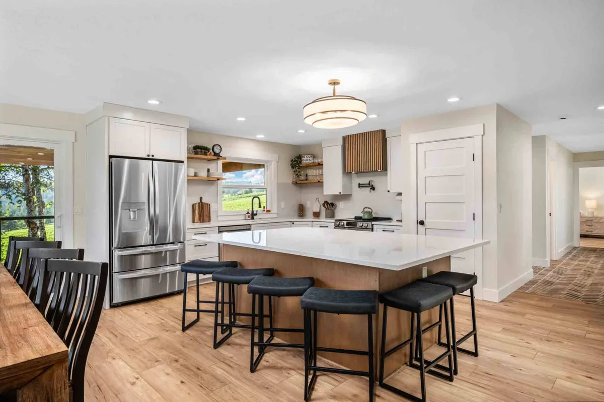 A spacious kitchen with a large island featuring comfortable seating, modern stainless steel appliances, and open wooden shelving. A wide window offers a glimpse of the outdoors, bathing the space in natural light. The kitchen is styled for entertaining and culinary creativity, with clean lines and designer touches.
