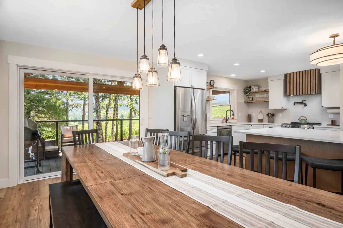 A warm, inviting dining area with a long wooden table set for a large group under elegant hanging light fixtures. The dining space flows seamlessly into the kitchen, and patio doors offer scenic views, suggesting a connection to the outdoors and the vineyards beyond.